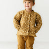 A young girl with a joyful expression, wearing an Organic Kids Wildflower organic merino wool buttoned jacket and mustard pants, stands against a white background.
