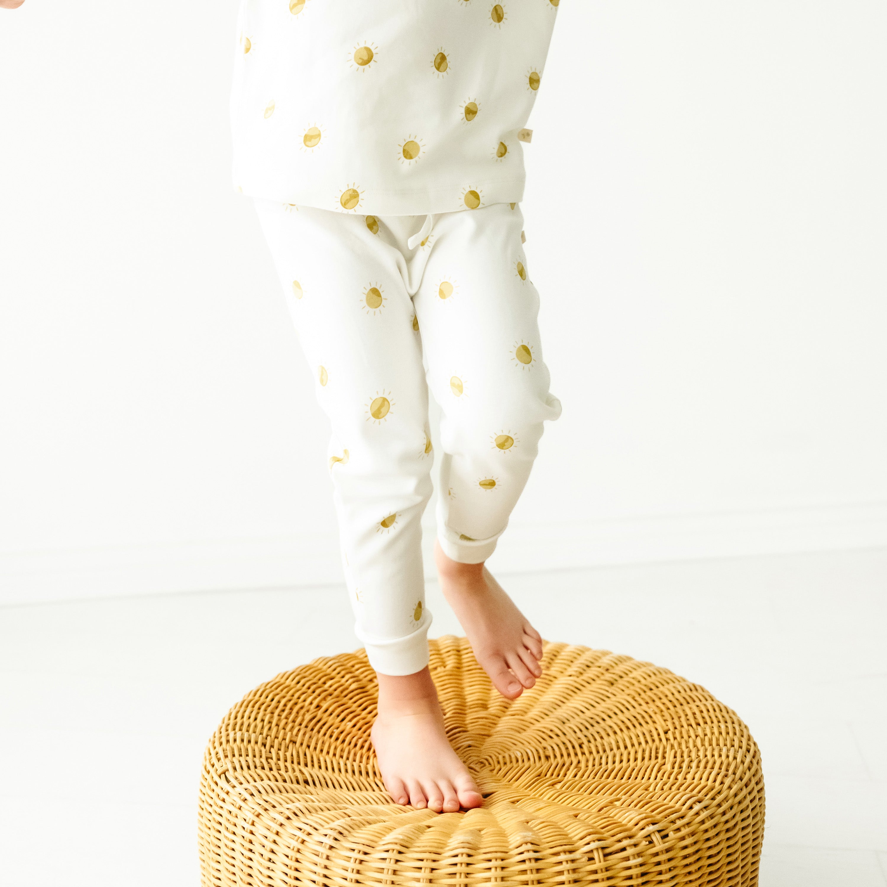 A baby in white pajamas with yellow sun prints stands on a wicker stool against a plain white background, only the lower half of their body visible wearing Organic Harem Pants - Sunshine by Makemake Organics.