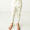 A toddler wearing Makemake Organics' Organic Harem Pants in Sunshine print, decorated with golden sun prints and a bow on the shirt, standing barefoot on a white floor against a white background.