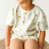A young child is seated on a wicker stool, wearing a Makemake Organics Organic Tee and Shorts Set - Sunshine adorned with sun motifs and matching shorts. The focus is on the outfit and the child's posture, with her face turned away.