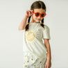 A young toddler in white clothing with a "good vibes" logo, wearing large round orange sunglasses and holding one of her braided pigtails, standing against a plain background in the Makemake Organics Boxy Tee and Skort Set - Sunshine.