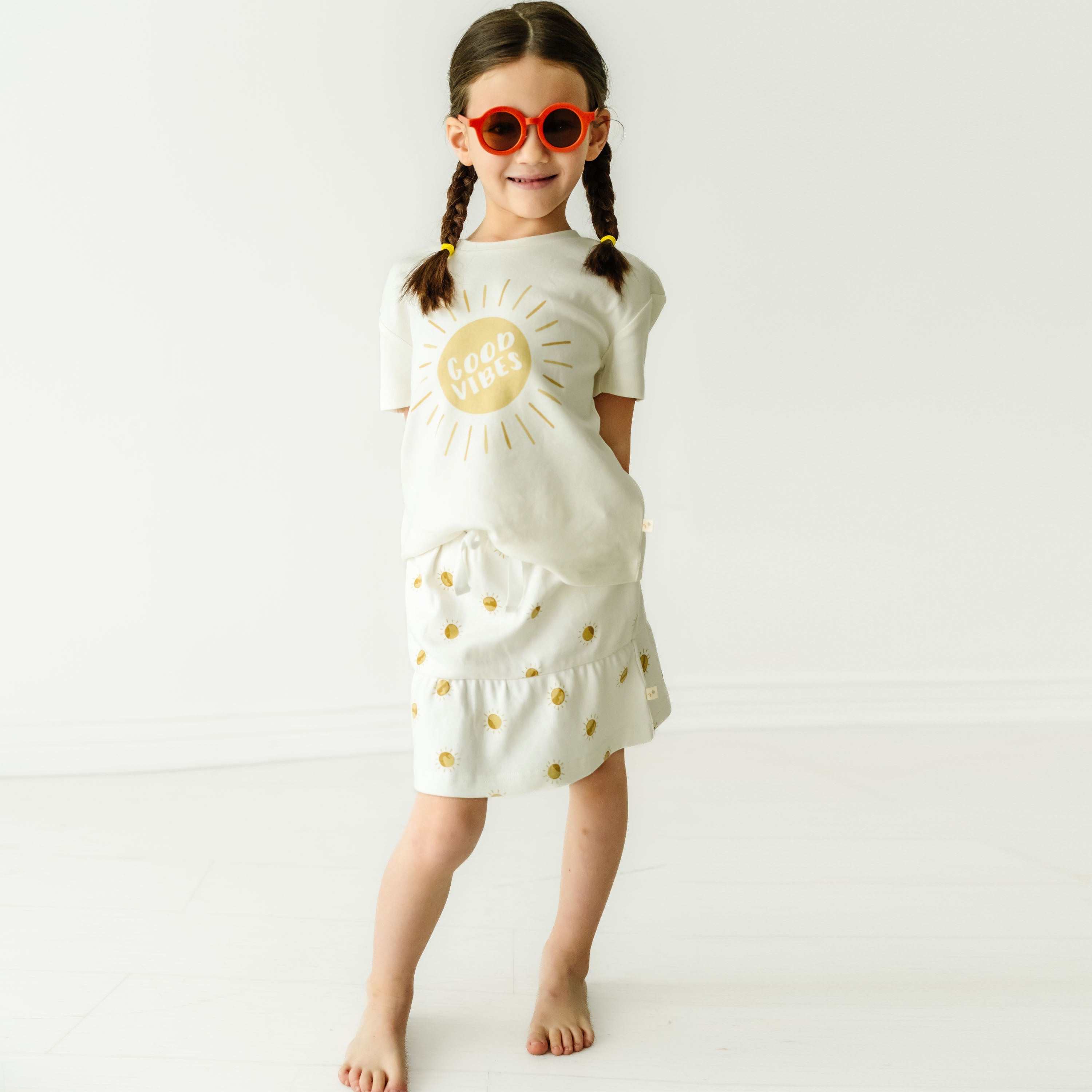 A young toddler with braided hair, wearing a Makemake Organics Boxy Tee and Skort Set - Sunshine, stands smiling in sunglasses against a plain background.