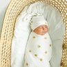 A newborn baby sleeps peacefully swaddled in a Makemake Organics Organic Swaddle Blanket & Hat - Sunshine, inside a woven basket on a light background.