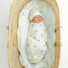 Newborn baby swaddled in a Makemake Organics Organic Swaddle Blanket & Hat - Sunshine, sleeping peacefully in a wicker bassinet, wearing a matching beanie.