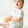 A baby in a Makemake Organics Organic Short Zip Romper - Sunshine sits on a white floor, holding a small yellow ball and looking upward with a curious expression.