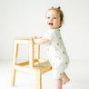 A happy baby with a top knot hairstyle stands next to a wooden chair in a bright room, wearing a Makemake Organics Organic Short Zip Romper - Sunshine with yellow floral patterns, smiling and looking away from the camera.