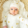 A toddler wearing a Makemake Organics Organic Kimono Knotted Sleep Gown - Sunshine, with sun designs and a matching hat, lying in a woven basket and looking up with wide blue eyes.