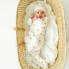 A baby in a white outfit with sun patterns lies in a woven, oval-shaped bassinet on a white background, looking upwards with one hand near the face, wearing the Organic Kimono Knotted Sleep Gown - Sunshine from Makemake Organics.