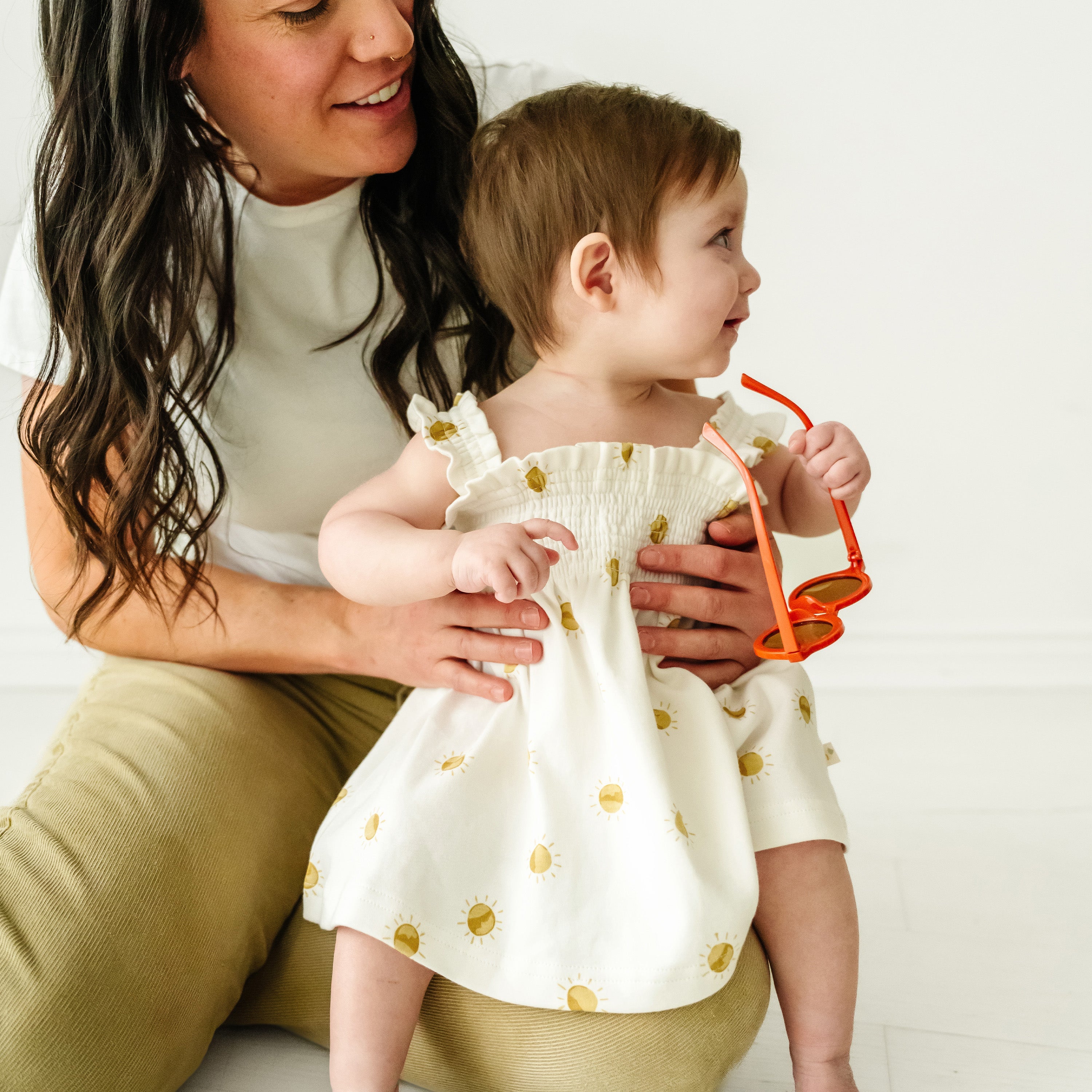 A woman smiling at a baby boy she's holding. The baby, wearing a Makemake Organics Organic Smocked Dress in Sunshine, looks to the side, holding red sunglasses. They're seated against a white background.