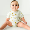 A happy toddler with a slight smile, wearing a Makemake Organics Organic Bubble Romper - Sunshine with yellow sun designs, sitting on a plain white background.