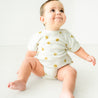 A happy toddler sits on a white background, wearing a Makemake Organics Organic Bubble Romper - Sunshine with yellow sun designs, looking up and to the side with a joyful expression.