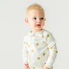 A baby with blond hair dressed in a Makemake Organics Organic 2-Way Zip Romper - Sunshine stands against a plain background, looking to the side with a curious expression.