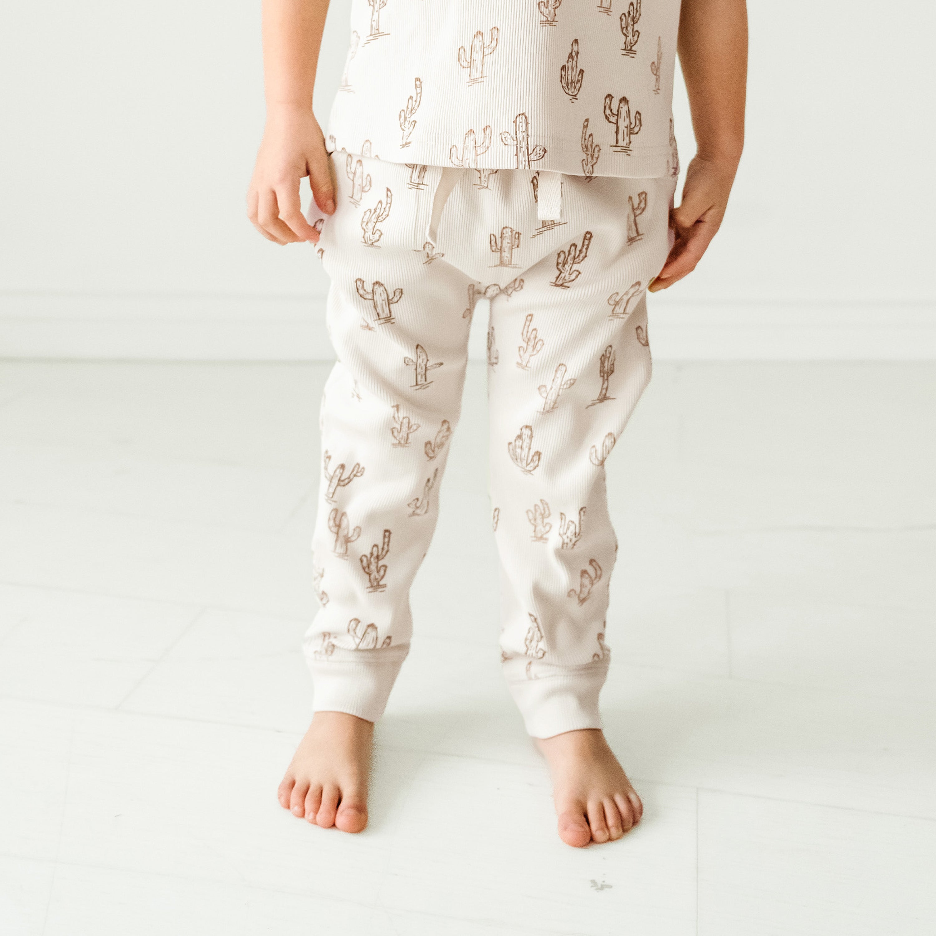 A baby wearing Makemake Organics' Organic Harem Pants in the Cactus pattern, including pants and a shirt with a ruffled hem, stands barefoot on a white floor against a white background.