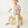 A toddler boy in a beige outfit with Makemake Organics Organic Short Sleeve Button Romper - Cactus print sits on a small wooden stool against a white background, looking surprised.