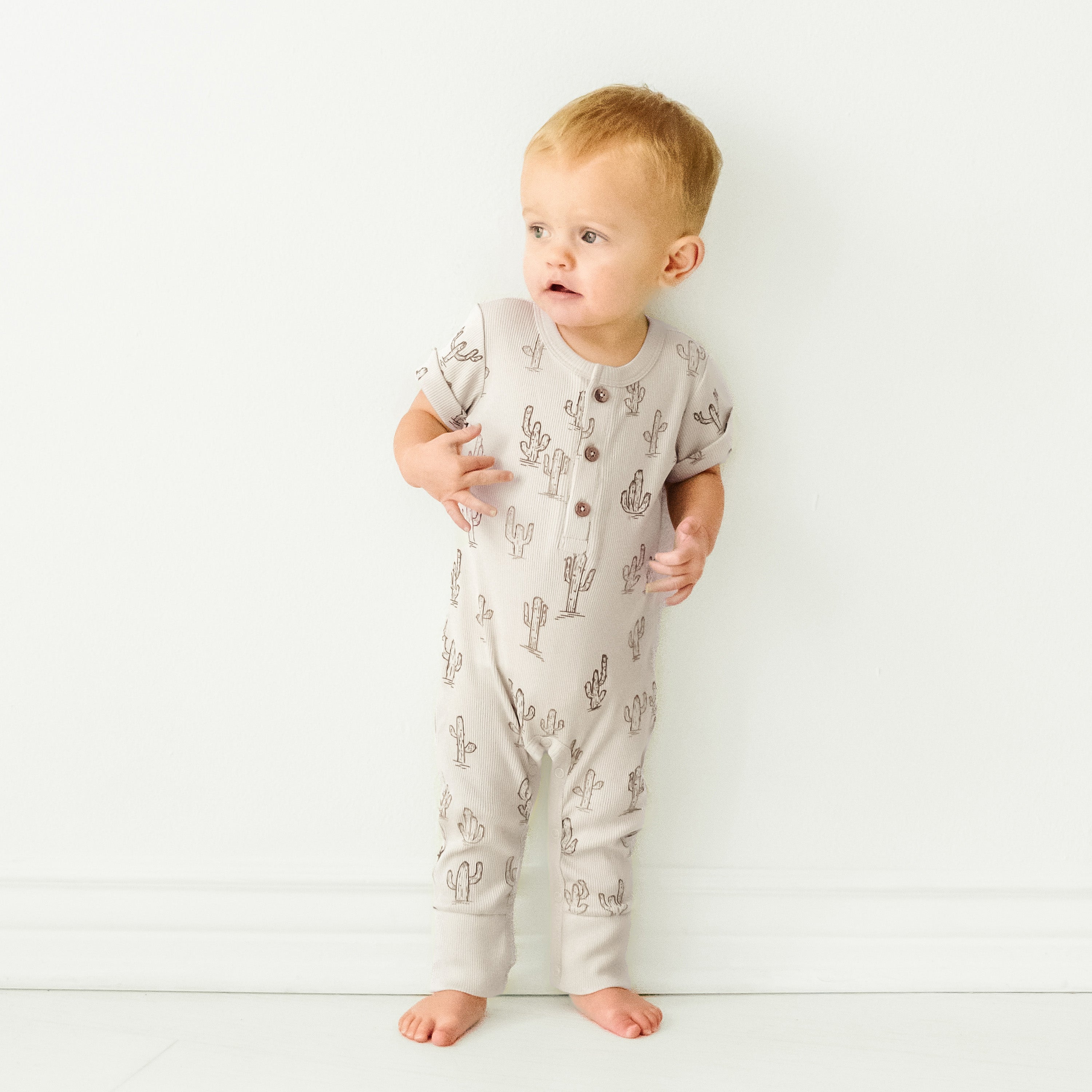 A baby with blond hair stands against a white wall, wearing a Makemake Organics Organic Short Sleeve Button Romper - Cactus, looking slightly to the side.