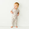 A toddler boy in a Makemake Organics cactus-print short sleeve button romper stands alone against a plain white background, looking to the side with a curious expression.