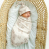 A newborn sleeps peacefully in a woven basket, swaddled in a Makemake Organics Organic Swaddle Blanket & Hat - Cactus set, on a soft white cushion.