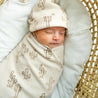 A newborn baby sleeps peacefully, swaddled in a Organic Swaddle Blanket & Hat - Cactus from Makemake Organics, inside a woven basket.