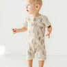 A baby in a beige Organic Short Zip Romper - Cactus from Makemake Organics stands in a bright, white room, looking to the side with a surprised expression.