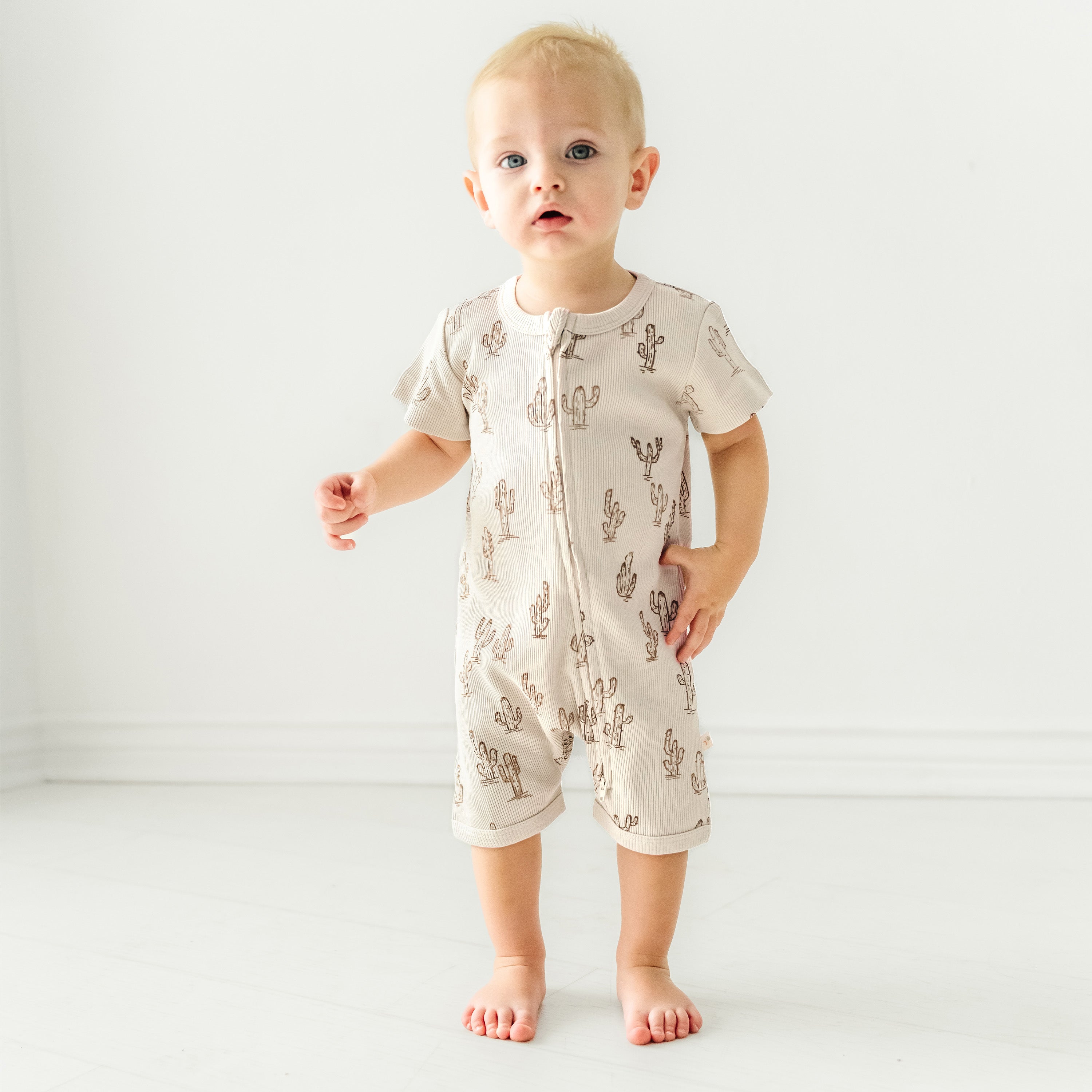 A toddler girl in a Makemake Organics Organic Short Zip Romper - Cactus standing barefoot on a white floor with a plain background, looking surprised.