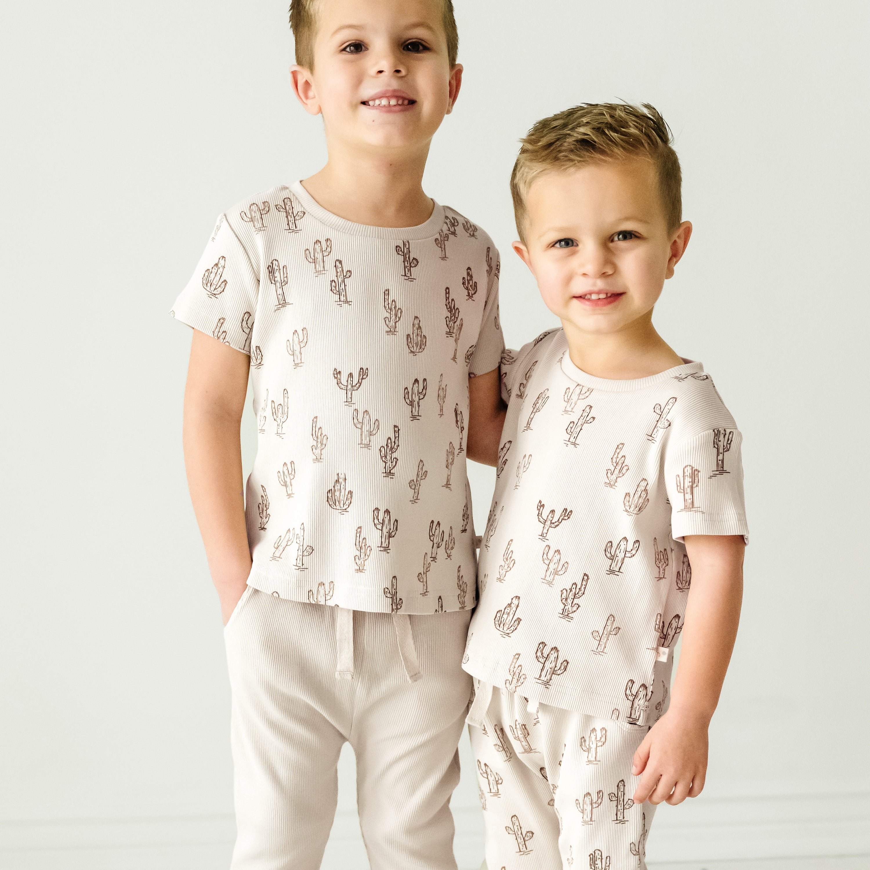 Two young boys smiling and standing together, one older and taller than the other. Both are wearing Makemake Organics Organic Tee & Pants Set - Cactus with a cactus print.