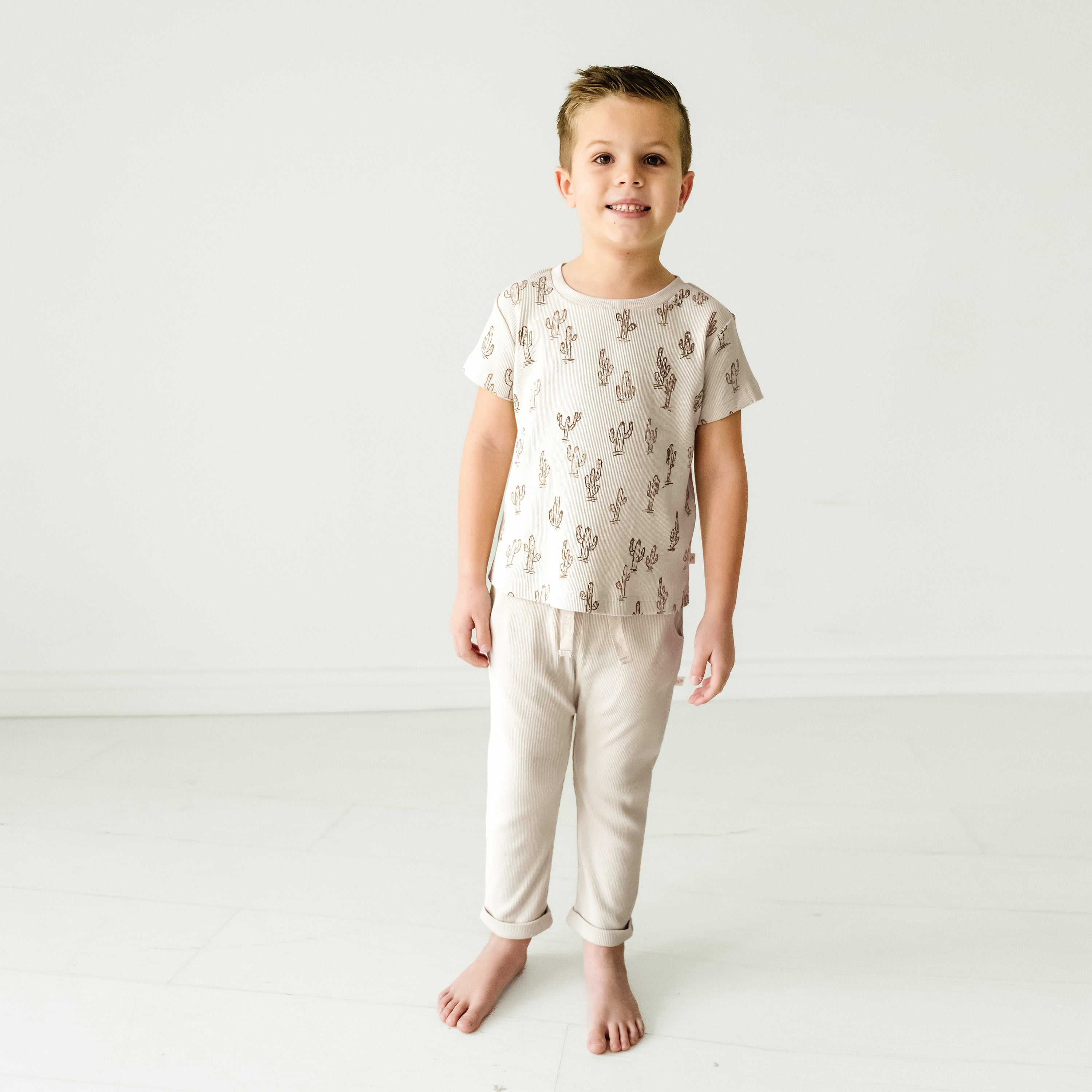 A young boy in a Makemake Organics Organic Tee & Pants Set - Cactus stands barefoot, smiling, in a plain white studio setting.