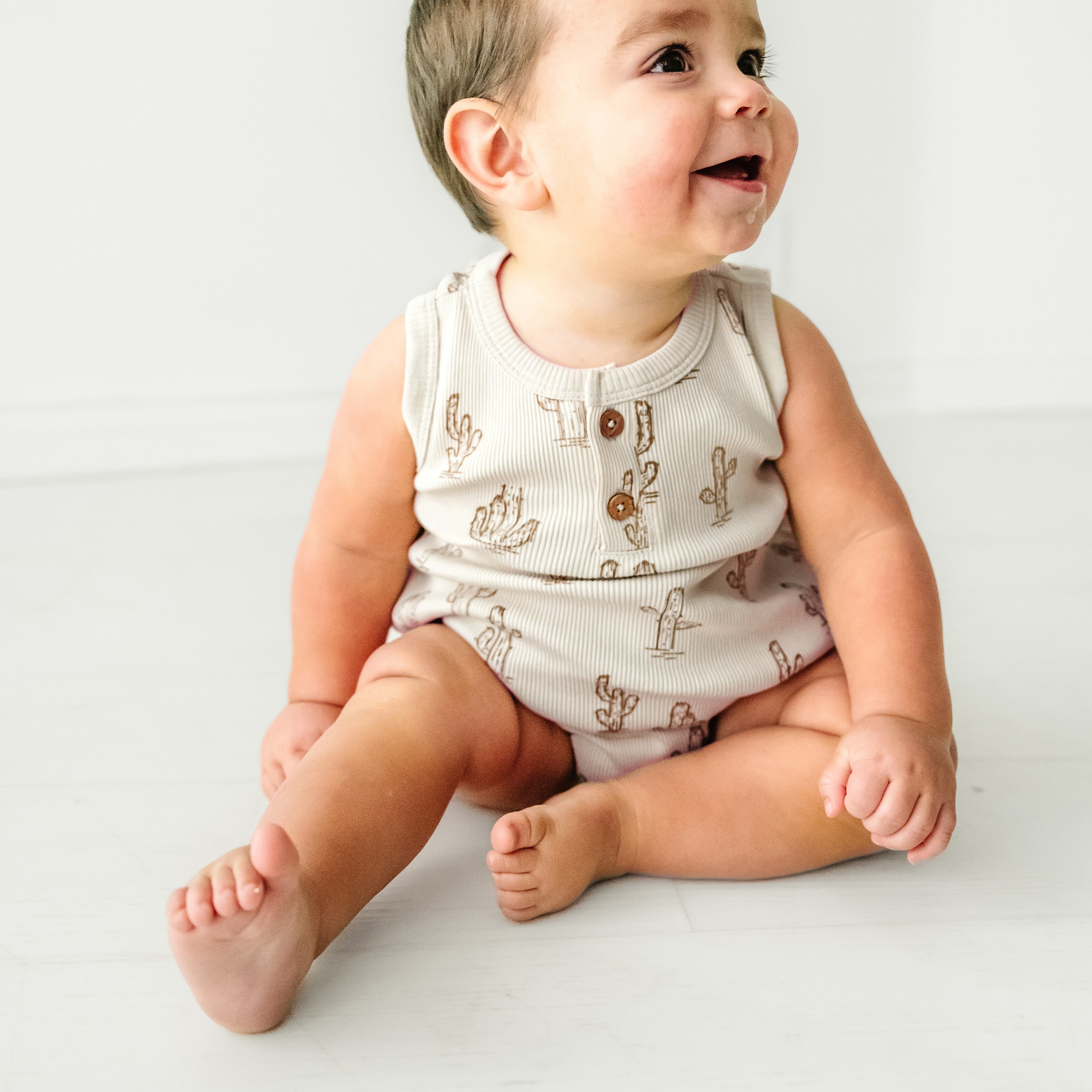 A joyful toddler in a Makemake Organics Organic Bubble Onesie - Cactus sits on a white floor, looking up and to the left with a big smile.