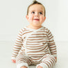 A joyful toddler with dark hair styled upwards, wearing a Makemake Organics Organic Kimono Top & Pants Set in Stripes, sitting on a white floor and looking up with wide, bright eyes and a happy expression.