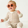 A toddler wearing a Makemake Organics Organic 2-Way Zip Romper in Stripes and orange sunglasses sits on a white background, looking cheerful yet slightly teary-eyed.