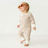 A smiling baby in a Makemake Organics Organic 2-Way Zip Romper - Stripes and orange sunglasses, standing unaided and appearing to take steps on a white background.