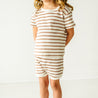 A young child wearing the Makemake Organics Organic Tee and Shorties Set in Stripes stands against a plain white background, looking down slightly, with their hands lightly touching.