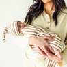 A woman holds a sleeping newborn baby swaddled in a Makemake Organics Organic Swaddle Blanket & Hat in Stripes. The background is plain, emphasizing the tender moment.