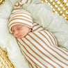 A newborn baby sleeps peacefully in a wicker basket, swaddled in a Makemake Organics Organic Swaddle Blanket & Hat - Stripes.
