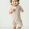 A joyful baby with curly hair smiling in a Makemake Organics Organic Short Zip Romper - Stripes, standing confidently in a brightly lit room.