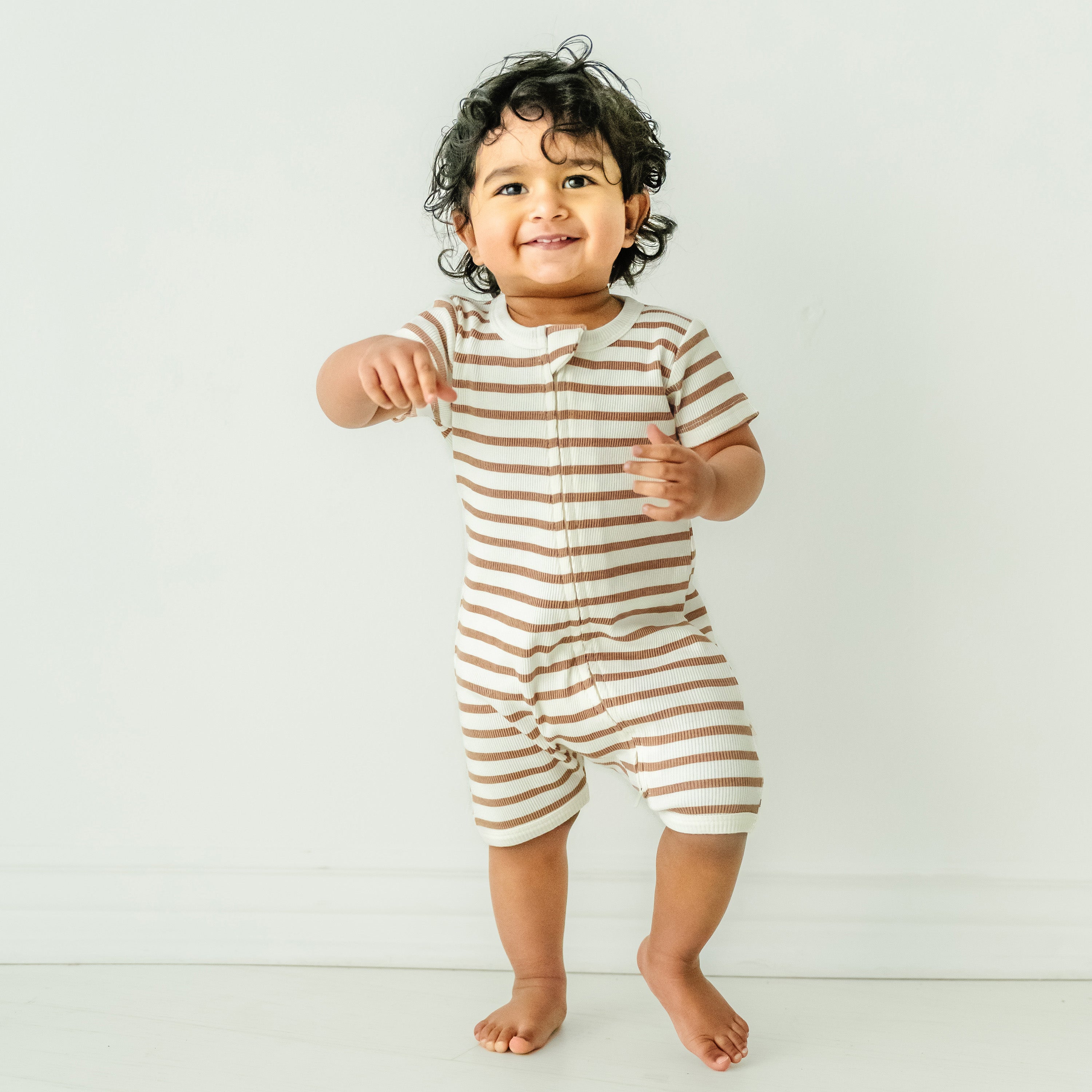 Toddler boy with curly hair, smiling and pointing, wearing a Makemake Organics Organic Short Zip Romper - Stripes against a plain white background.