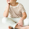 A young child sits on the floor, wearing a Makemake Organics Organic Tee & Pants Set - Stripes, resting their chin on their hand with a thoughtful expression.