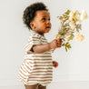 A baby with curly hair, dressed in a Makemake Organics Organic Bubble Romper - Stripes, holds a bouquet of flowers, looking to the side with a curious expression, in a well-lit room with a white background.