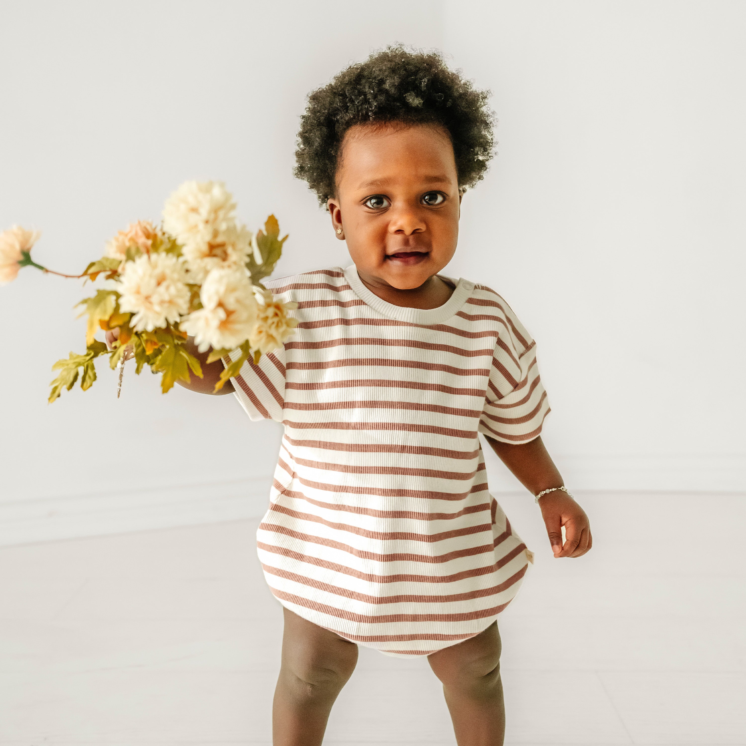 A baby with curly hair, dressed in a Makemake Organics Organic Bubble Romper - Stripes, happily holding a bouquet of cream-colored flowers, standing in a bright room with a white background.