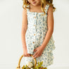 A toddler girl in a Makemake Organics Organic Muslin Peplum Top and Shorts Set - Periwinkle holding a small wicker basket filled with flowers, standing against a plain light background.
