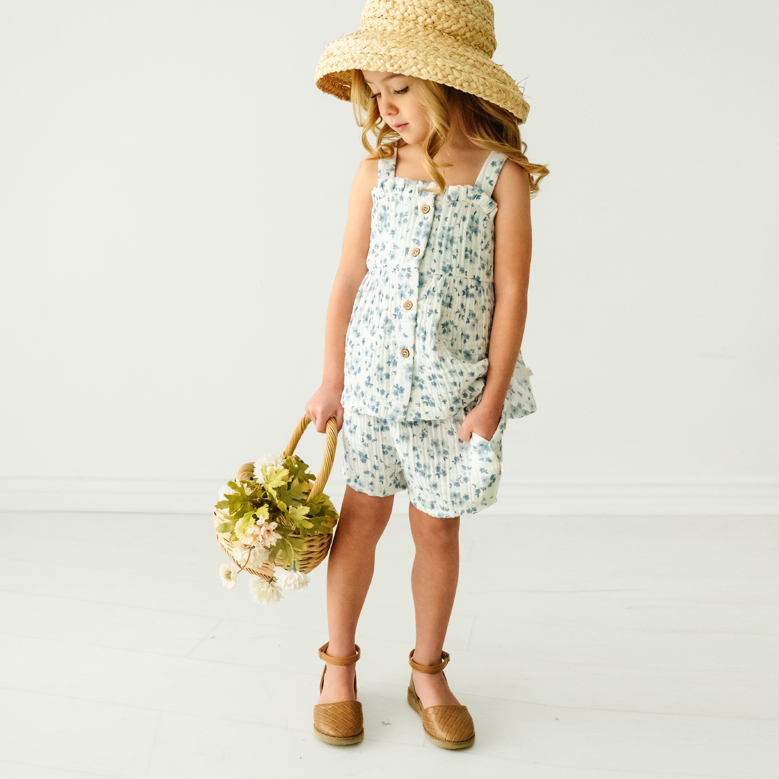 A young girl in a Makemake Organics Organic Muslin Peplum Top and Shorts Set - Periwinkle and straw hat holds a basket of flowers, standing in a minimalist white studio space.
