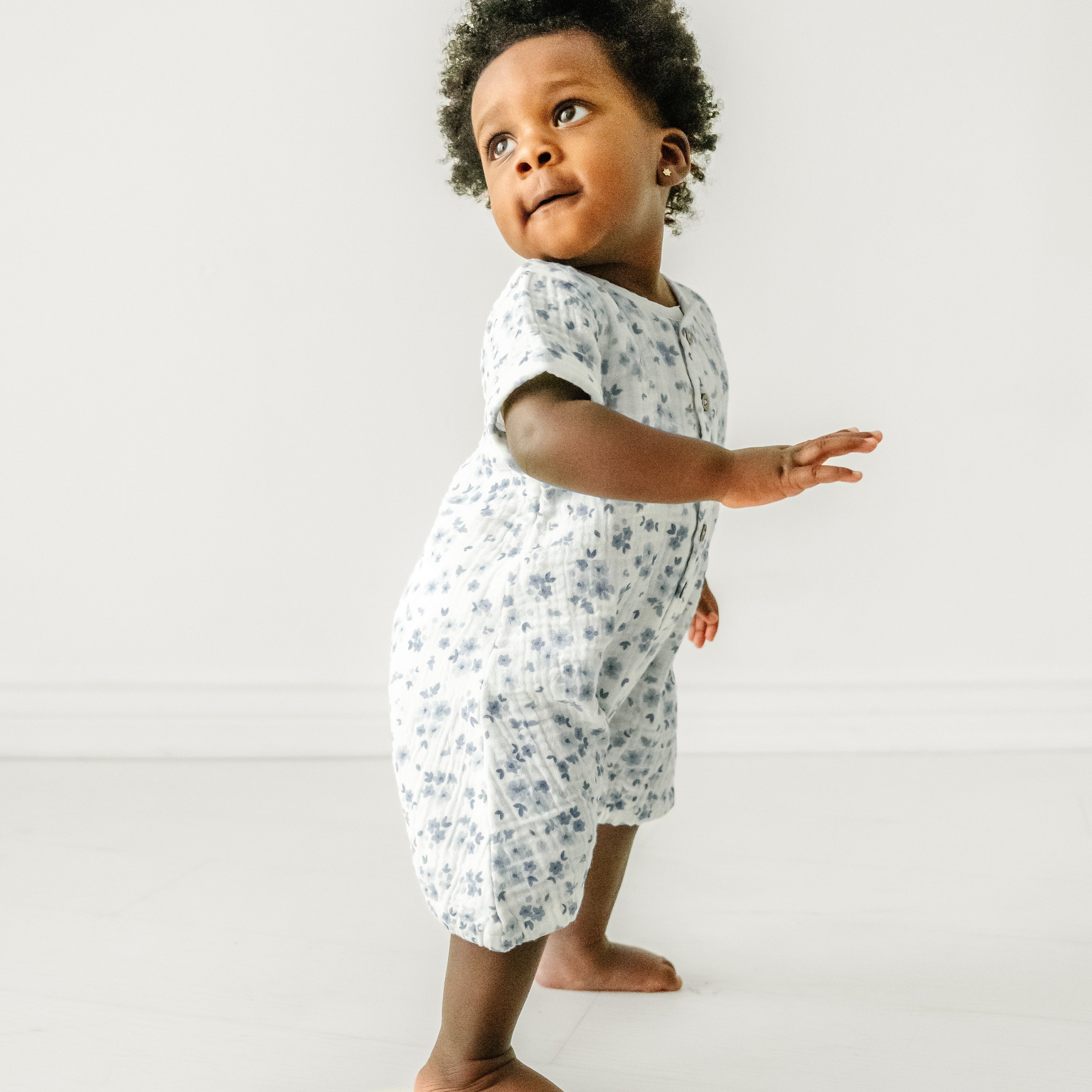 A young toddler girl taking tentative steps in a Makemake Organics Organic Muslin Short Bubble Romper in Periwinkle, against a plain white background, looks joyfully to the side while stretching out a hand.