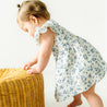 A toddler girl in a Makemake Organics Organic Muslin Button Flutter Dress - Periwinkle stands, leaning on a woven wicker basket in a bright room, appearing curious and focused.