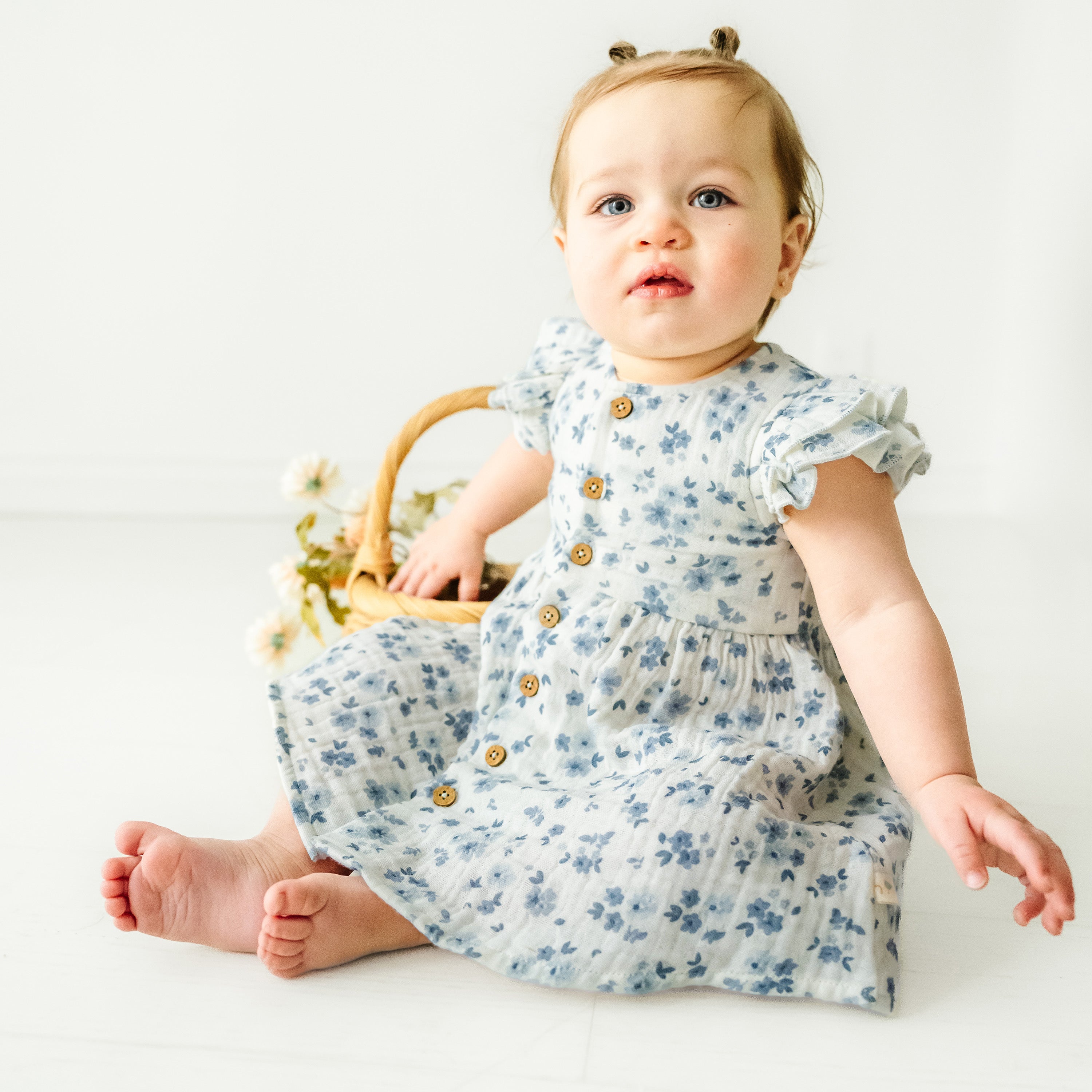 A baby girl with light brown hair in pigtails, wearing a Makemake Organics Organic Muslin Button Flutter Dress in Periwinkle, sits on a white floor next to a basket of flowers, looking curious.