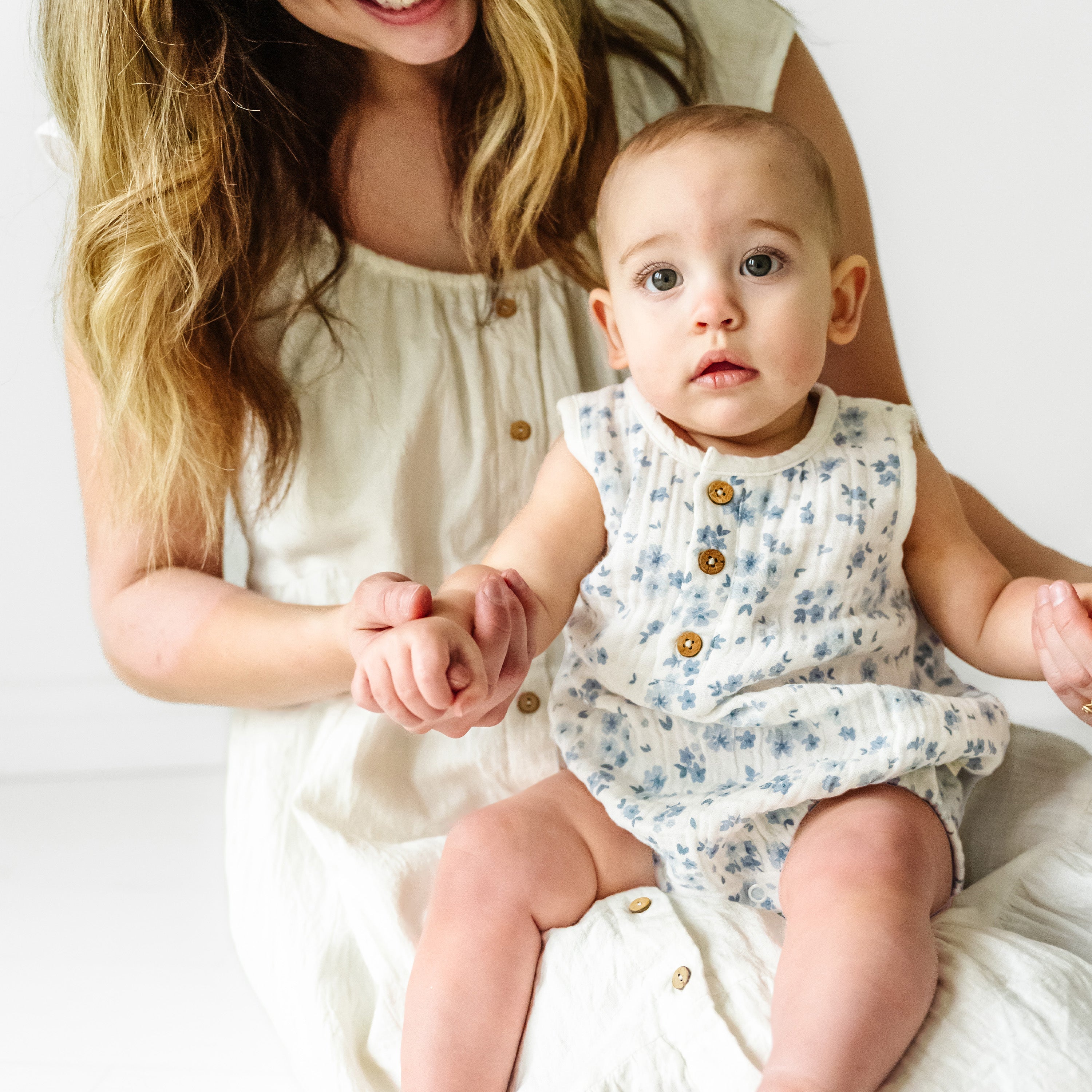 A woman with long, curly blonde hair holding hands with a toddler girl in a floral dress, both sitting against a white background.