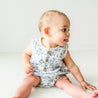 A toddler in a Makemake Organics Organic Muslin Bubble Onesie in Periwinkle sits on the floor, sticking out their tongue playfully while glancing to the side.