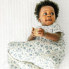 A toddler with curly hair, wearing a floral outfit, lies on a white quilt, looking upwards with a curious expression while holding a small toy and wearing the Muslin Wearable Blanket in Periwinkle by Makemake Organics.