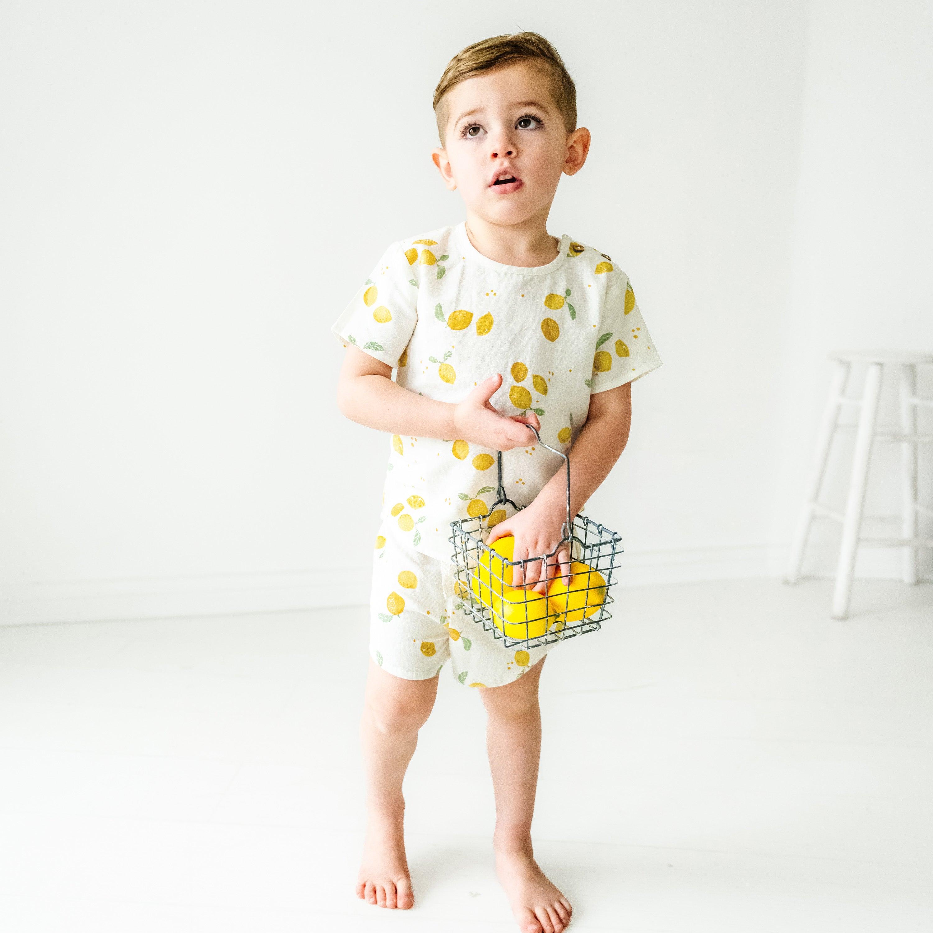 A young girl in a white outfit with lemon prints stands barefoot, holding a small yellow wire basket, looking slightly surprised in a bright, white room wearing the Organic Linen Top and Shorts 2 Piece Set in Citron by Makemake Organics.