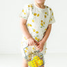 A young toddler wearing Makemake Organics' Organic Linen Top and Shorts 2 Piece Set - Citron holds a small metal basket containing lemons, standing against a plain white background.