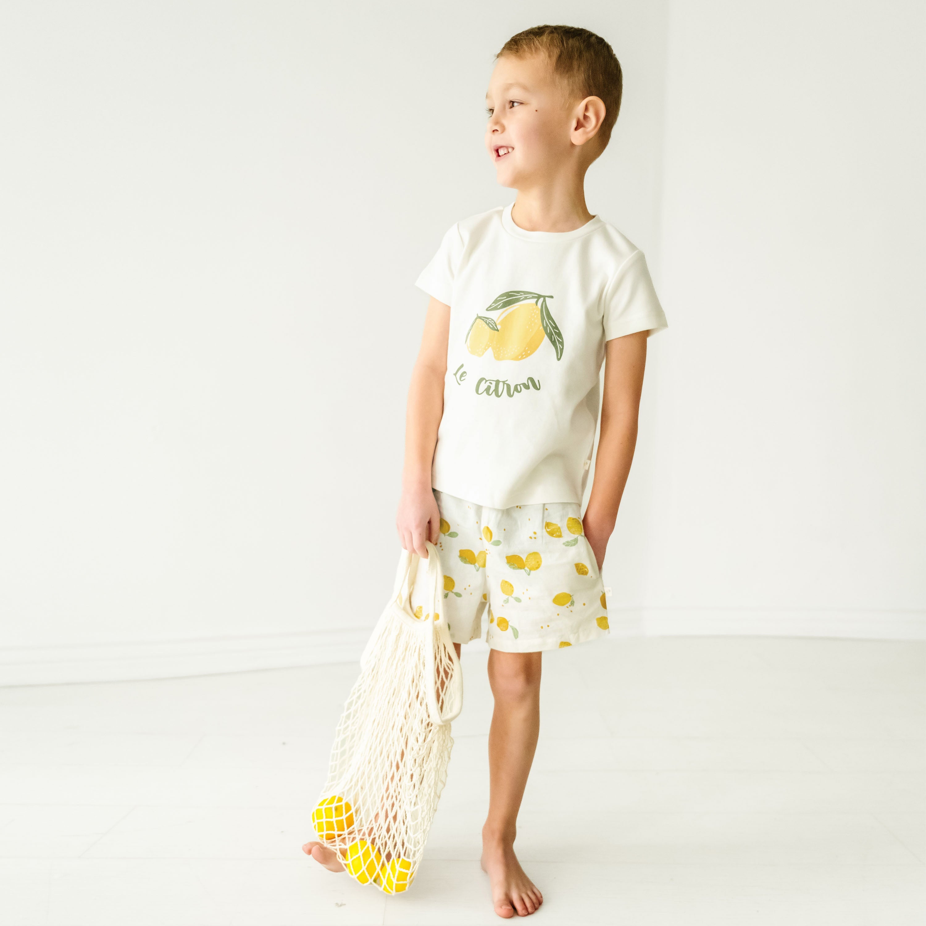A toddler in a Organic Crew Neck Tee - Le Citron t-shirt and lemon-print shorts smiles while holding a net bag filled with lemons, standing in a bright room with a white background.