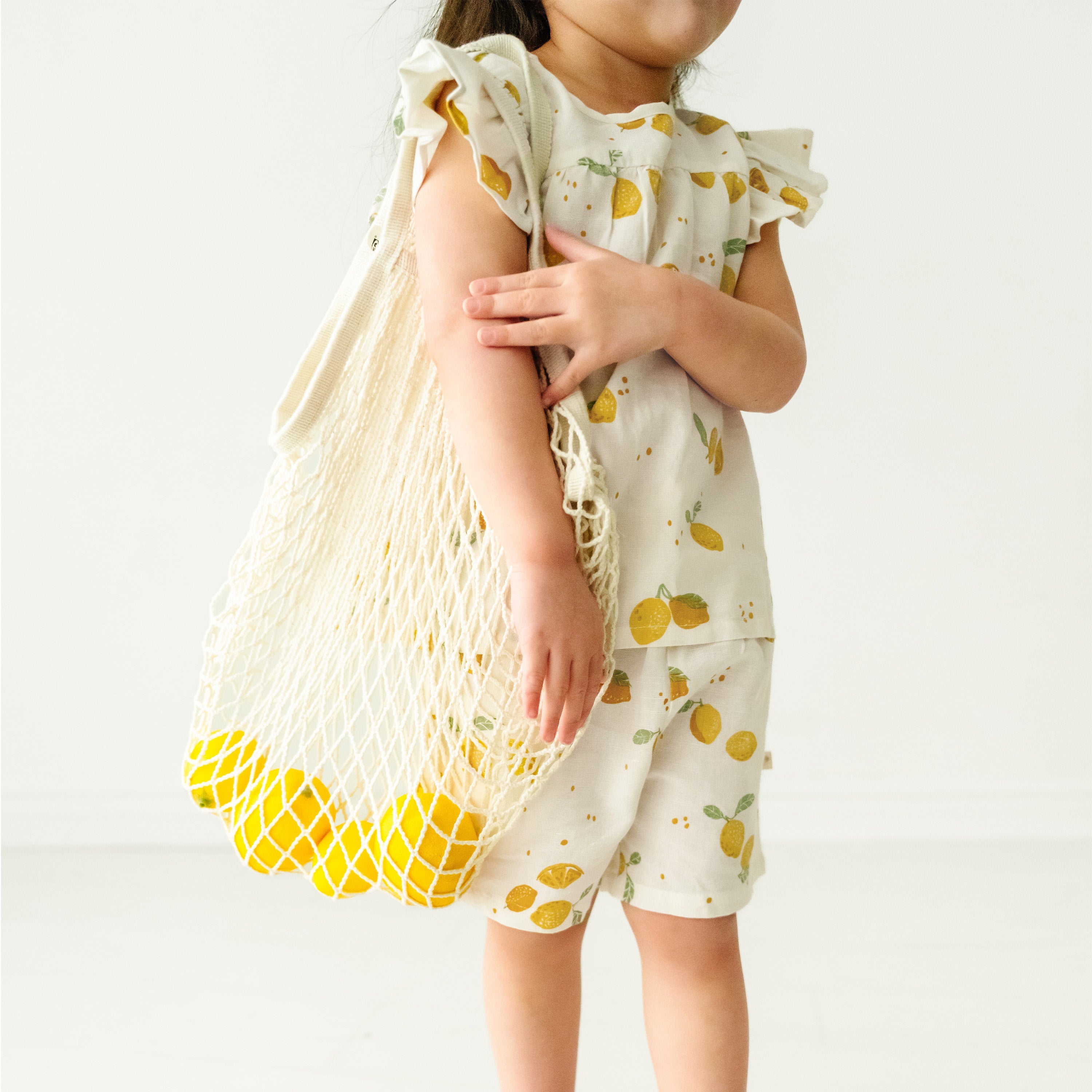 A young toddler in a Citron Organic Linen Flutter Top and Shorts from Makemake Organics holds a mesh bag full of lemons, standing against a plain background. The focus is on the bag and the toddler's arm.